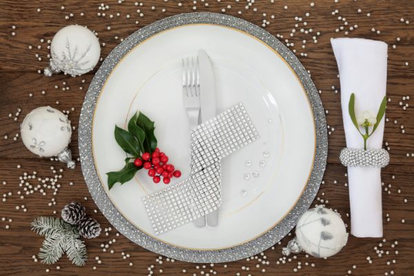Christmas dinner place setting with plate, napkin, cutlery, bauble decorations with holly and mistletoe over oak table background.