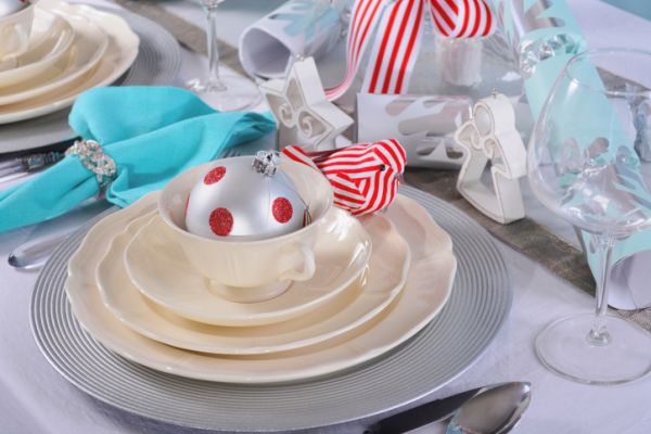 Modern Christmas table place setting with fine china and silver charger plates on white tablecloth and white tree branch centerpiece with stripe birds against a pale blue backdrop, closeup.